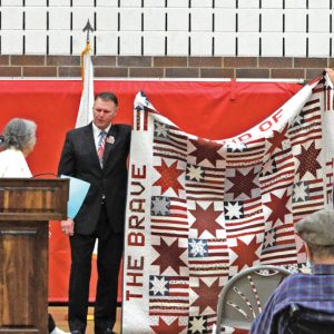 QUILT OF VALOR — Sally Johnson of Colfax presented a Quilt of Valor she had made to William C. Yingst Jr., school district superintendent, during the Veterans Day program at Colfax High School November 11. Johnson’s husband, Glen (Pete) Johnson, was a Vietnam veteran who died in an accident in Minneapolis in 2011. Johnson said she did not know how she was going to get through her grief, so she started making Quilts of Valor that she presents to veterans. Johnson said she makes two of every quilt, one to keep for Pete and one to give away to a veteran. The Quilt of Valor was a complete surprise to Yingst, who is a retired Command Sergeant Major with the United States Army. Eric Johnson, the son of Sally and Pete, is on the right, holding up a corner of the quilt. —photo by Rich Meredith
